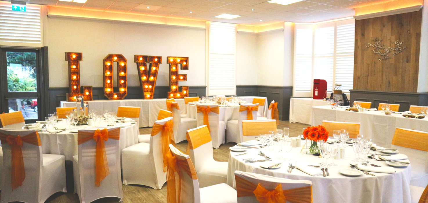 Wedding reception with round tables, white linens, orange chair sashes, floral centerpieces and illuminated 'LOVE' marquee.