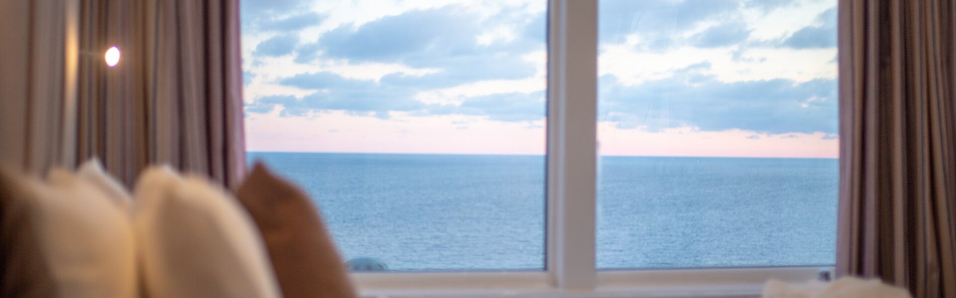 Ocean-view guest room with plush pillows and large window framing a pastel sunset over the sea, soft curtains at sides.