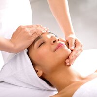 Guest receiving a relaxing facial in the hotel spa; therapist's hands on forehead and jaw, towel-wrapped head, peaceful expression