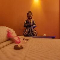 Bronze Buddha statue under warm lamp beside folded towels, pink flower petals and massage stones on a spa table.