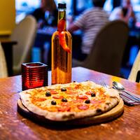 Wood-fired pepperoni pizza with black olives on wooden board, spicy chili-infused oil bottle and cozy restaurant table.