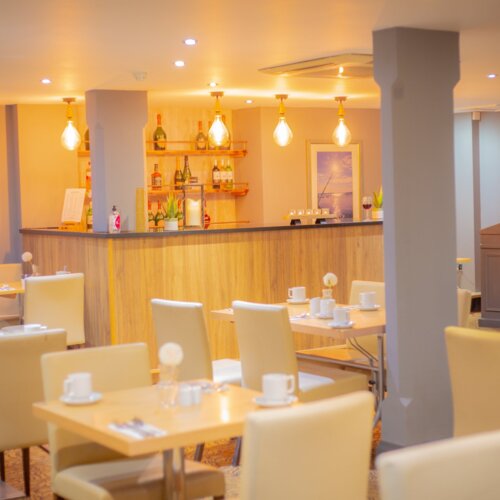 Warm hotel dining room with wooden bar, pendant bulbs, wine shelves and cream leather chairs set for coffee.