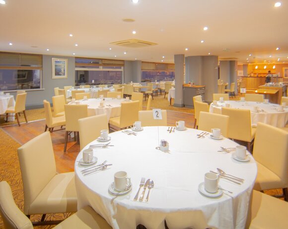Spacious hotel dining room with round white-clothed tables, beige chairs, place settings and a buffet under warm lighting.