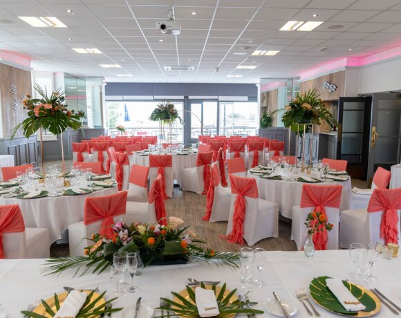 Waterfront banquet room set for wedding with round tables, white chair covers, coral sashes and tropical floral centerpieces