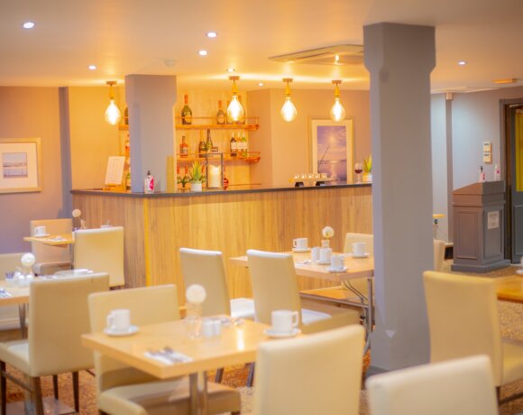 Warm hotel dining room with wooden bar, pendant bulbs, wine shelves and cream leather chairs set for coffee.