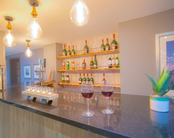 Hotel bar with dark stone counter, hanging Edison bulbs, two glasses of red wine, back shelves of bottles and cozy lighting