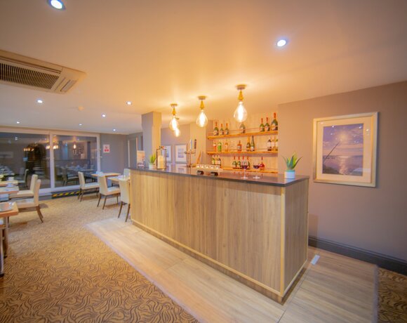Cozy hotel bar and dining area with wooden counter, exposed pendant bulbs, wall shelves of spirits and table seating.