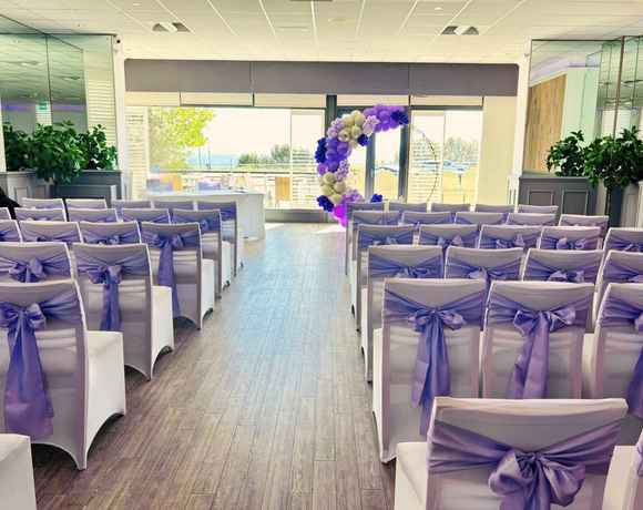 Bright waterfront ceremony room with rows of white chair covers tied with lavender bows, circular balloon arch by glass doors and outdoor terrace view.