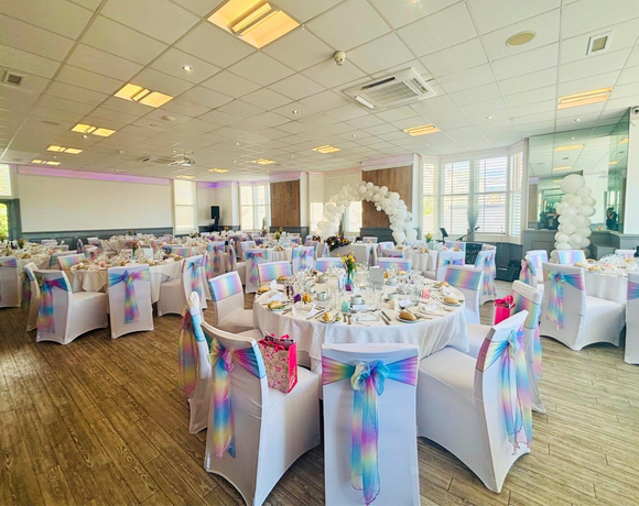 Bright banquet room set for a wedding with round tables, white chairs with pastel rainbow sashes and a balloon arch