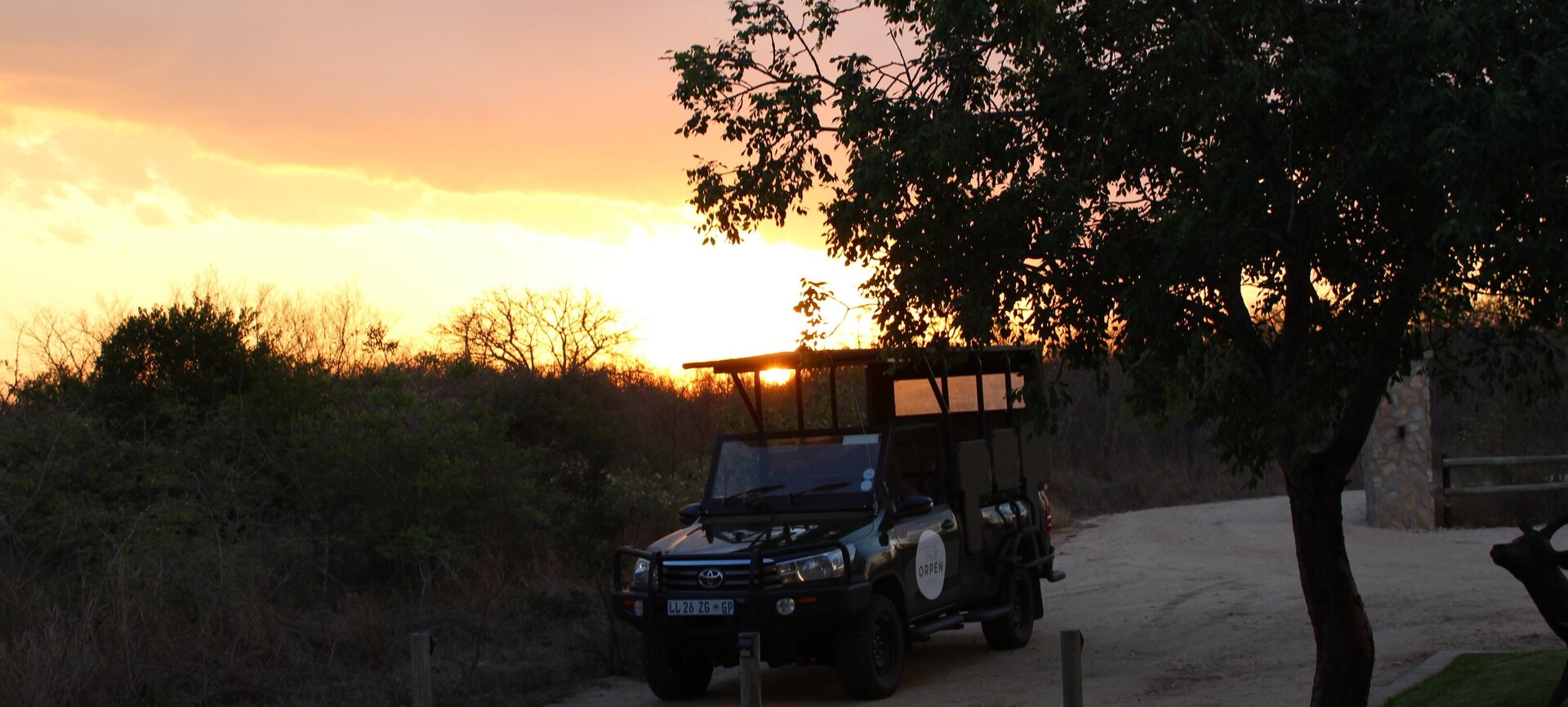 Open-top safari vehicle parked on a lodge driveway beneath trees, golden sunset over scrubby bush, serene evening