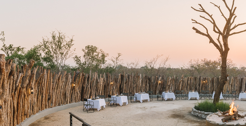 Walled open-air dining courtyard with white-clothed tables, lanterns, central fire pit and sunset over bushveld.