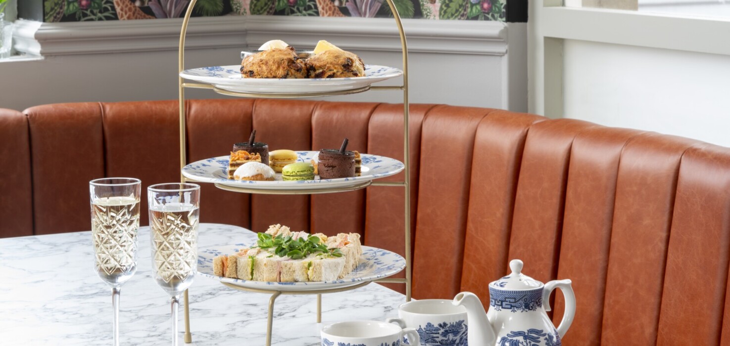 Three-tier afternoon tea with scones and finger sandwiches on blue-and-white porcelain, marble table and leather banquette.
