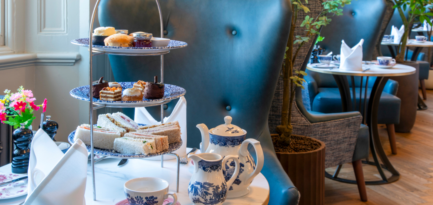 Three-tier afternoon tea with finger sandwiches, scones and pastries, blue-and-white teapot on marble table in a tea lounge.