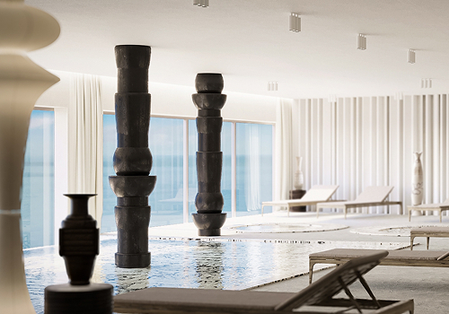 Minimalistischer Innen-Spa-Pool mit skulpturalen dunklen Säulen, Liegen und bodentiefen Fenstern mit Blick auf das ruhige Meer.