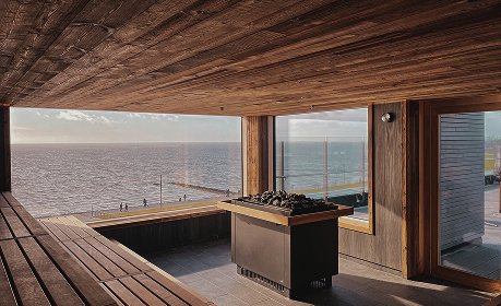 Salon spa wykończony drewnem z ławkami, centralnym paleniskiem i panoramicznymi oknami z widokiem na spokojny taras nad morzem.