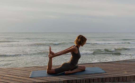 Gast macht Yoga auf hölzerner Strandterrasse bei Sonnenaufgang, dehnt sich auf Matte, ruhiges Meer mit sanften Wellen