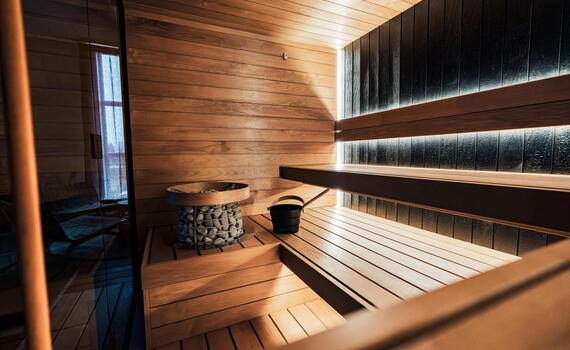Nowoczesna sauna wyłożona drewnem, piętrowe ławki, piec z kamieni, wiaderko, szklane drzwi i delikatne podświetlenie