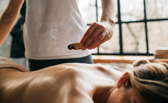 Terapeutka wykonuje relaksujący masaż pleców, trzymając dymiący ziołowy patyczek nad leżącym gościem przy dużym oknie.