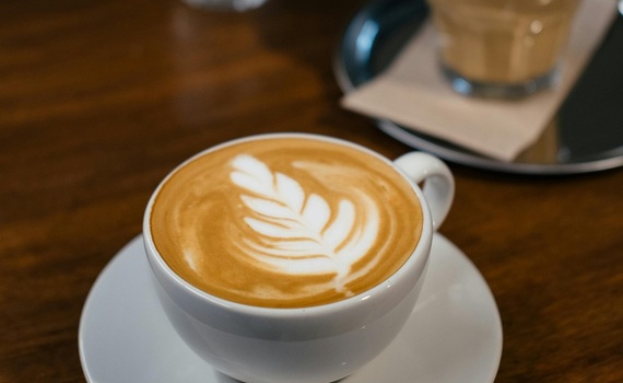 Cappuccino mit Blatt-Latteart in weißer Tasse auf warmem Holztisch, zweite Latte und kleine Vase im Hintergrund unscharf
