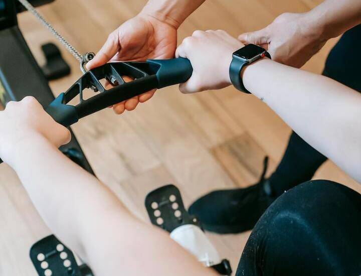 Trener hotelowej siłowni pomaga gościowi na maszynie wioślarskiej, zbliżenie dłoni, pasy na stopach i smartwatch na drewnianej podłodze