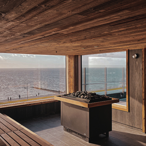 Sauna z drewnianym wnętrzem, piecem z kamieniami i ławkami, z panoramicznym widokiem na plażę i morze.