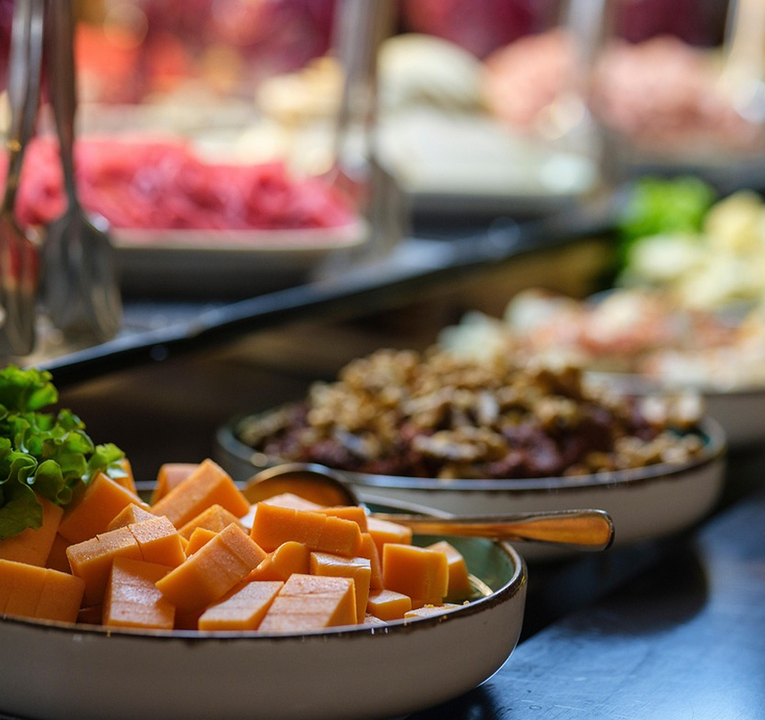 Kostki cheddaru i sałata na półmisku bufetowym, w tle nieostre wędliny, sałatki i łyżki do serwowania