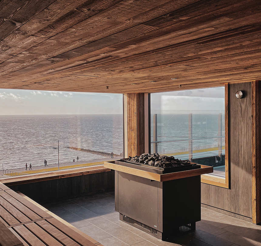 Sauna z drewnianym wnętrzem, panoramicznymi oknami na morze, ławkami i piecem z kamieni, widok na nadmorski deptak