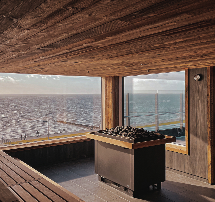 Holzsauna mit Panoramablick aufs Meer, mehrstufige Bänke und zentraler Steinofen; Menschen spazieren auf der sonnigen Promenade