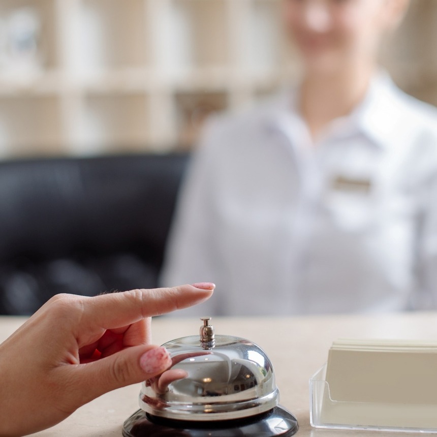 Gość naciska polerowany dzwonek na recepcji hotelu, recepcjonistka w mundurze rozmyta w tle.