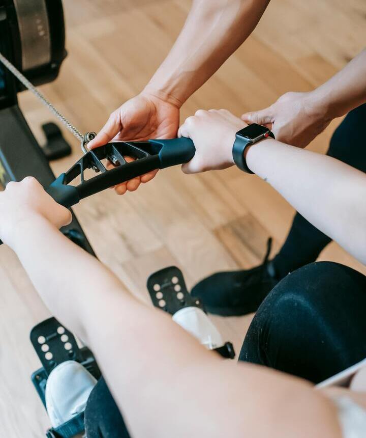 Trener hotelowej siłowni pomaga gościowi na maszynie wioślarskiej, zbliżenie dłoni, pasy na stopach i smartwatch na drewnianej podłodze