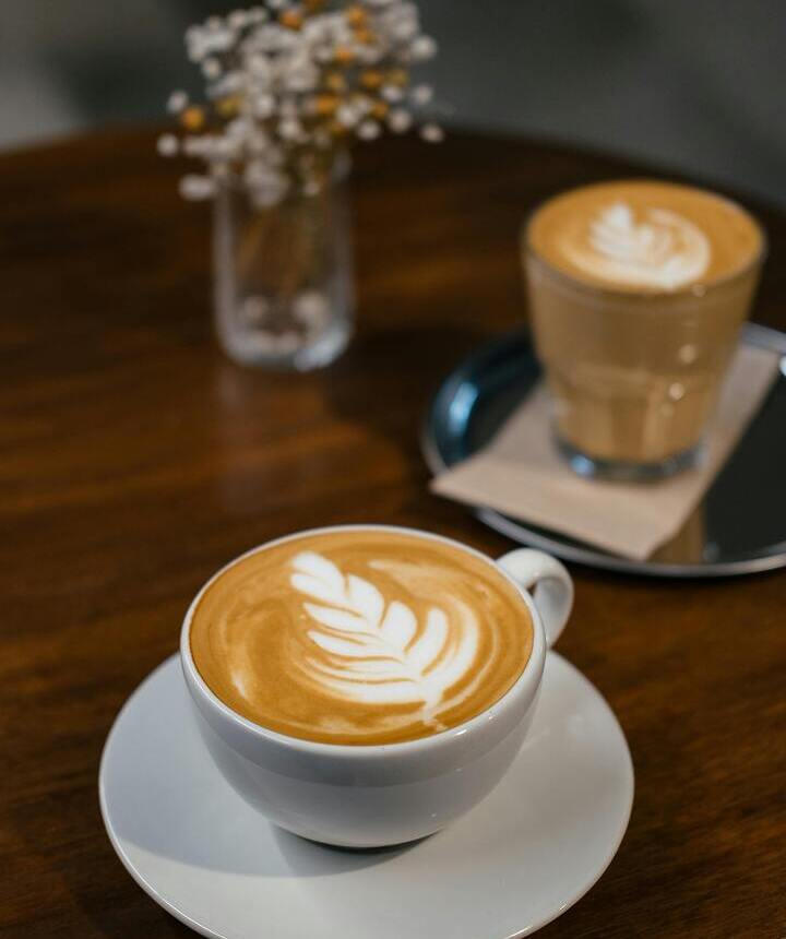 Dwie latte z ornamentem listka na drewnianym stoliku — biała filiżanka na spodku i szklanka na tacy obok małego wazonika z suszonymi kwiatami.