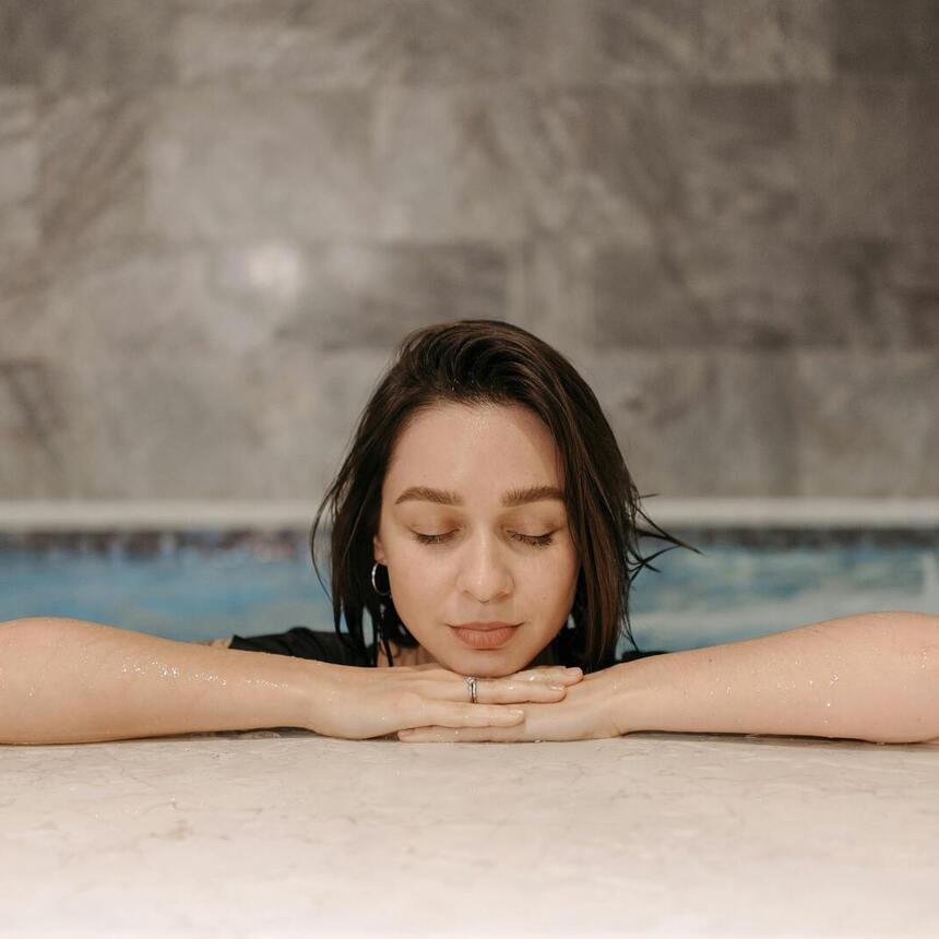 Gast entspannt am marmorumsäumten Innen-Spa-Pool, stützt das Kinn auf die Hände neben einem sanften Wasserfall und ruhigem blauem Wasser.