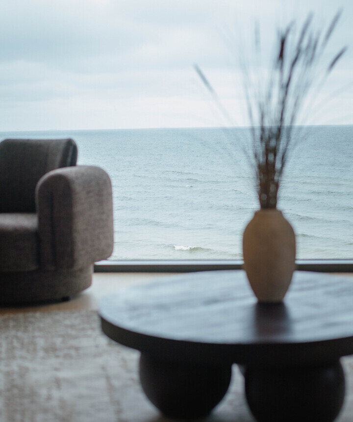 Lounge mit Sessel, rundem Holztisch und Vase, bodentiefes Panoramafenster mit Blick auf das ruhige Meer.