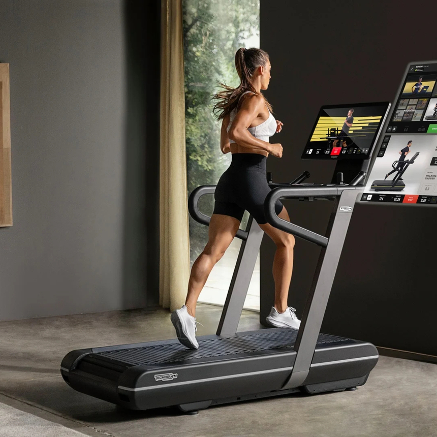Gast läuft auf modernem Laufband mit großen interaktiven Displays, die virtuelle Strecken und Trainings im hellen Hotel-Fitnessbereich zeigen.
