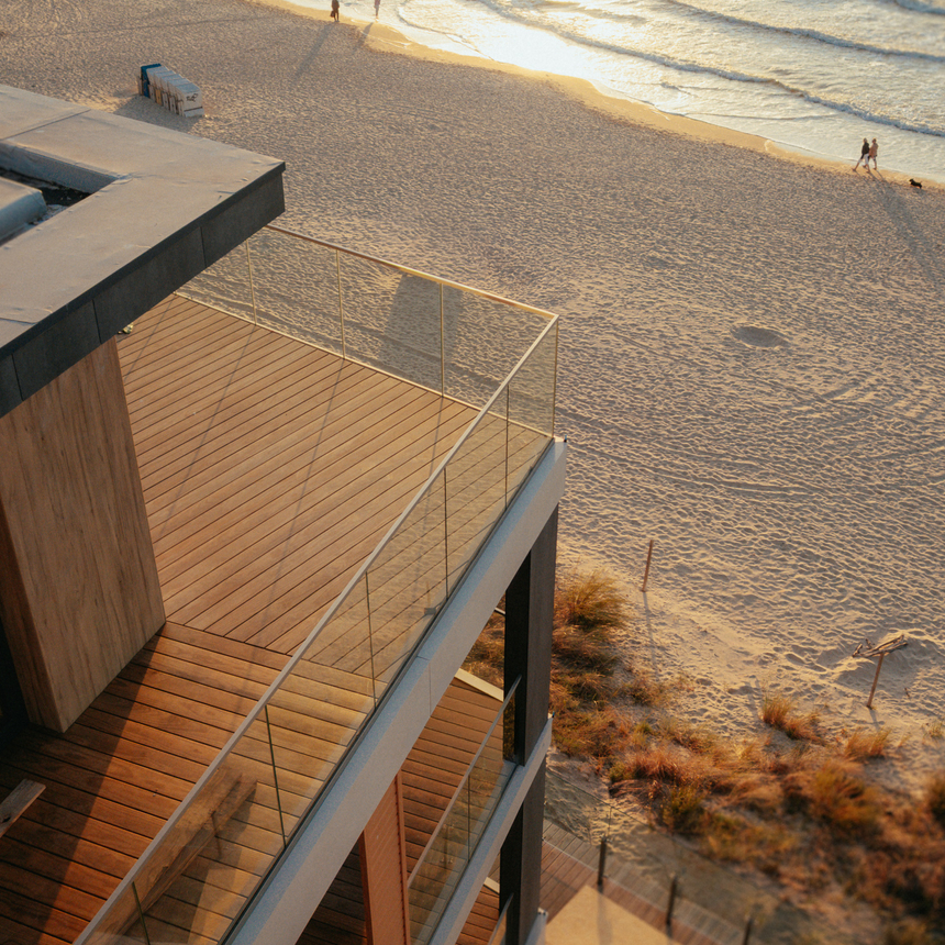 Nowoczesny apartament z drewnianym tarasem i szklaną balustradą nad piaszczystą plażą, falami i parą oglądającą zachód słońca.