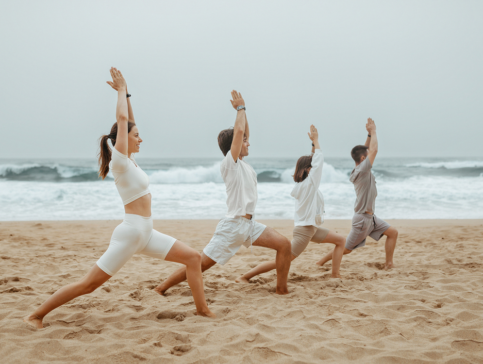 Cztery osoby w jasnym stroju ćwiczą jogę na piaszczystej plaży (poza wojownika) przy falach oceanu