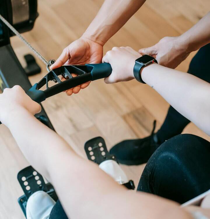 Trener hotelowej siłowni pomaga gościowi na maszynie wioślarskiej, zbliżenie dłoni, pasy na stopach i smartwatch na drewnianej podłodze