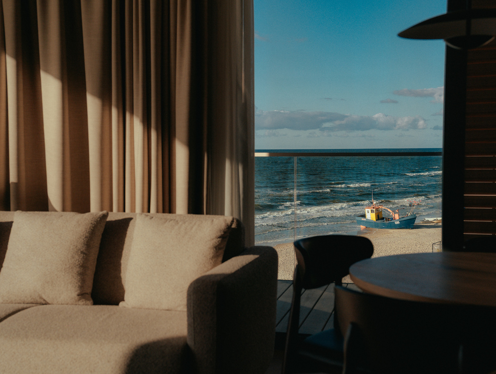 Przytulny salon w apartamencie z kanapą i zasłonami, balkon z widokiem na plażę, morze i małą łódź rybacką