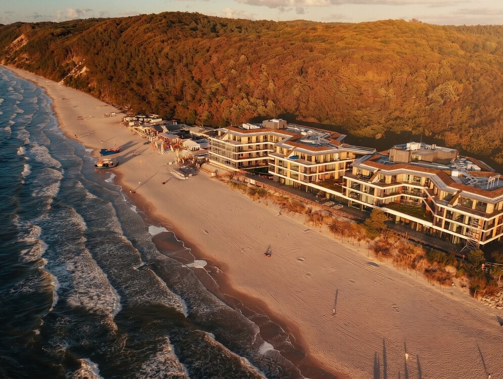 Nowoczesny hotel przy plaży z tarasowymi balkonami, barami i łodziami na szerokiej piaszczystej plaży u zalesionych wydm w świetle zachodu słońca.