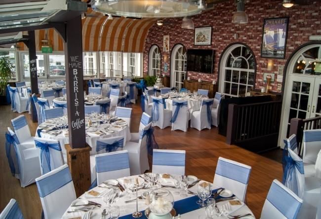 Hotel banquet room with round tables, white linens, navy chair sashes, glassware and centerpieces beneath arched windows.