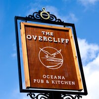 Wooden hotel pub sign 'The Overcliff — Oceana Pub & Kitchen' on ornate iron frame against bright blue sky