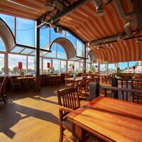 Sunlit seaside dining room with orange-striped vaulted ceiling, arched windows and wooden tables facing ocean views.