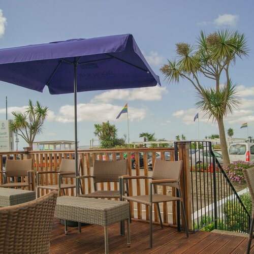 Coastal hotel terrace with wicker bar seating under a large purple umbrella, palm trees, flowerbeds and ocean glimpses beyond.