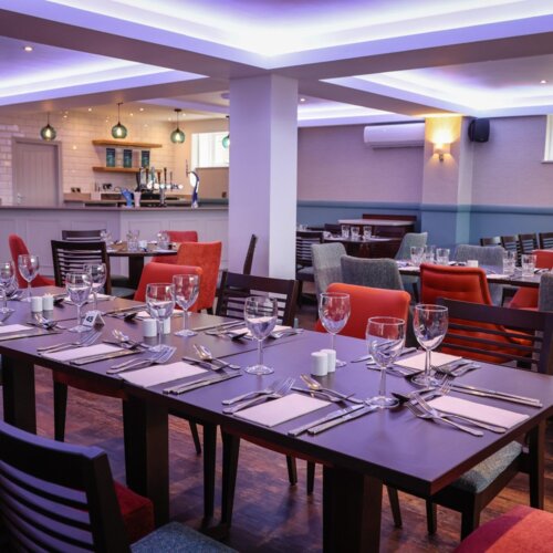 Hotel dining room with set tables, wine glasses, mixed upholstered chairs and back bar under soft pendant and ceiling lighting.