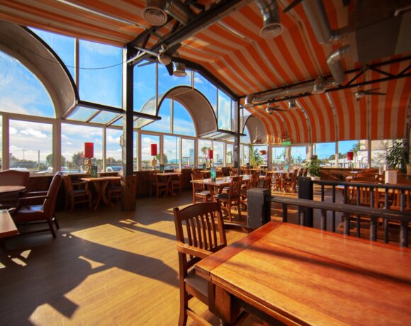 Sunlit seaside dining room with orange-striped vaulted ceiling, arched windows and wooden tables facing ocean views.
