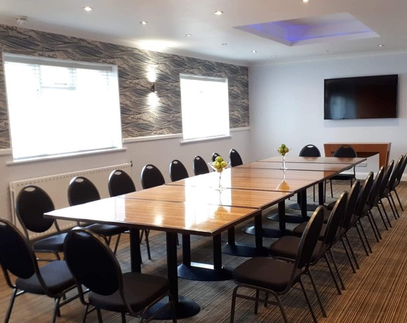 Hotel meeting room with long joined wood tables, black chairs, wall-mounted TV, blue-lit ceiling and glass vases with apples.