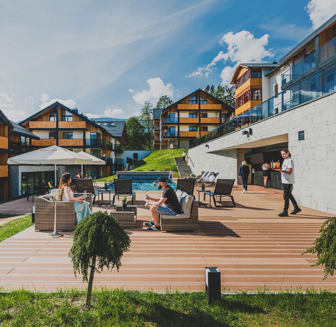 Taras przy basenie z leżakami i rattanowymi fotelami, barman podaje napoje, nowoczesne drewniane budynki hotelu w górach.