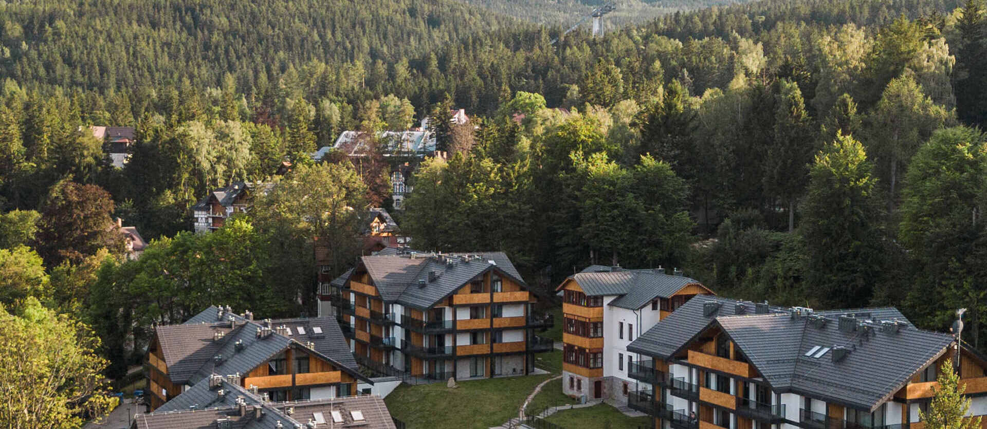 Górski resort w stylu chalet z balkonami, odkrytym basenem i widokiem na zalesioną dolinę i szczyty pod błękitnym niebem.