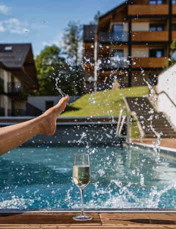 Zewnętrzny basen hotelowy, gość pluska nogą, kieliszek szampana na drewnianym tarasie, słoneczne balkony w stylu chalet