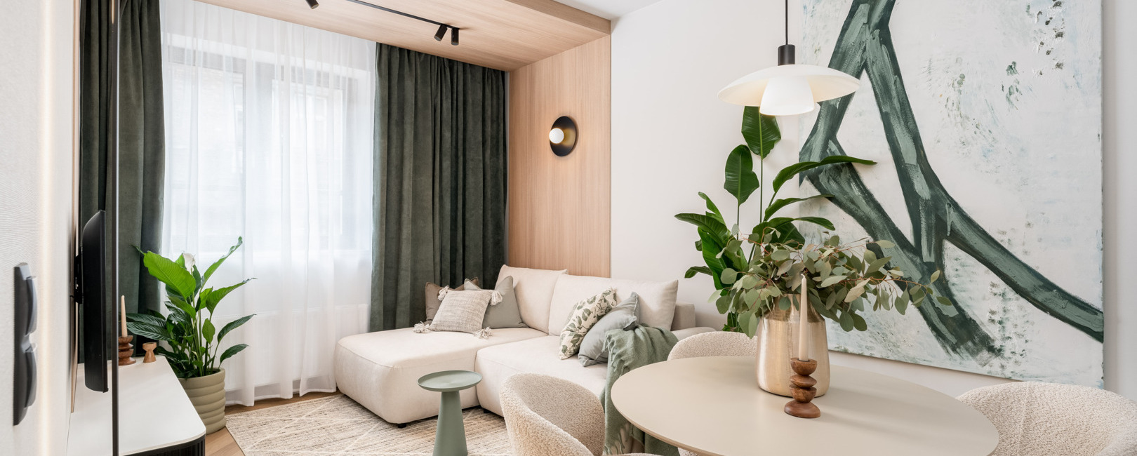 Nowoczesny salon hotelowy z beżową sofą narożną, okrągłym stołem i miękkimi fotelami w towarzystwie roślin i dużego obrazu.